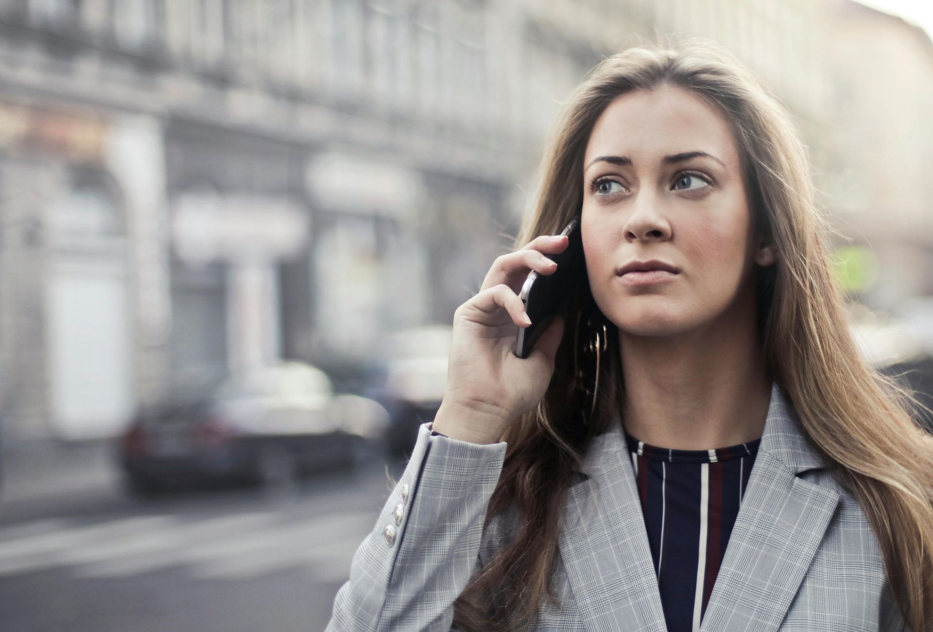 Woman in a gray blazer talking on a phone outdoors, city street in background.