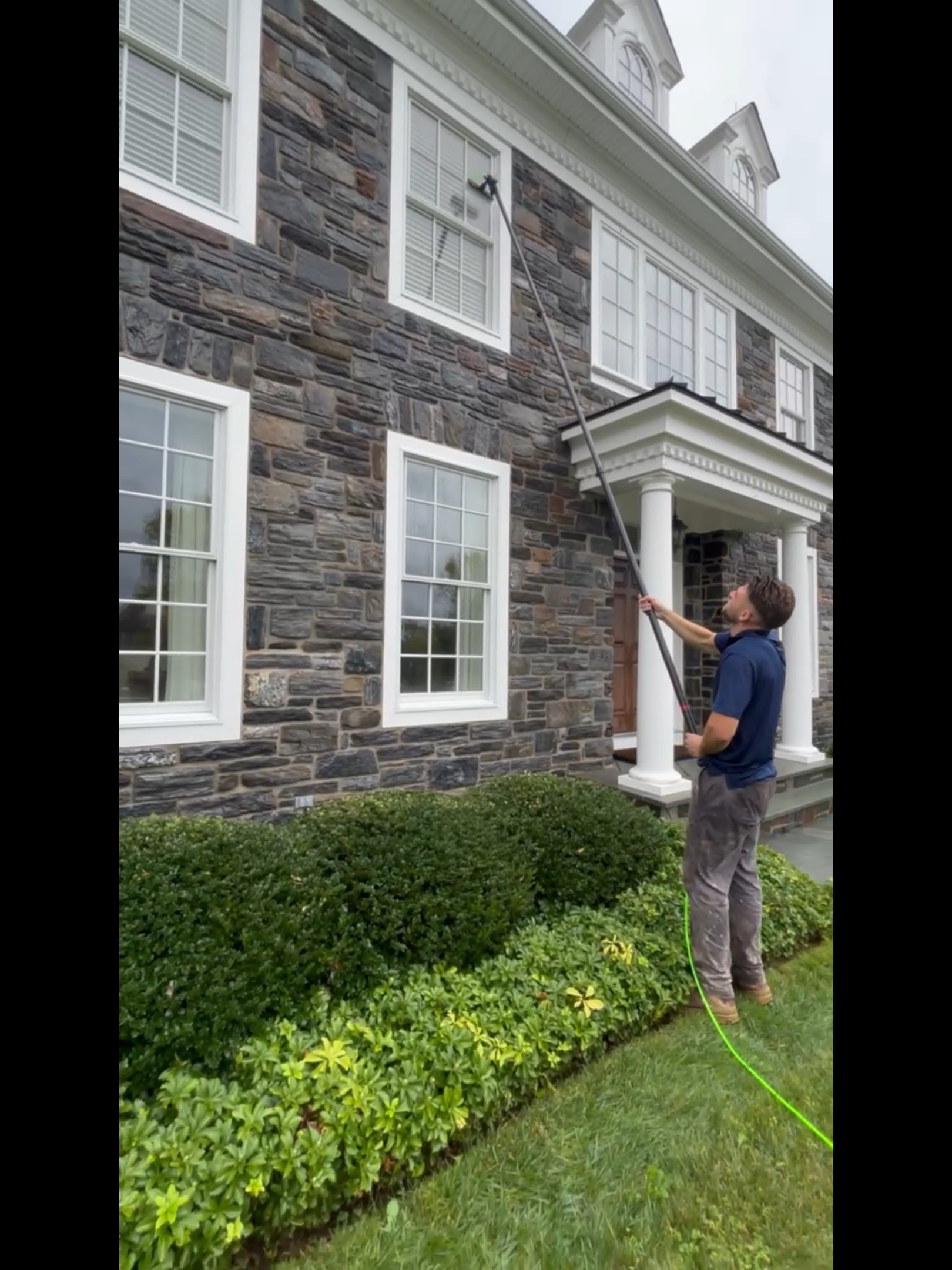 A man is cleaning the windows of a large stone house with a hose.