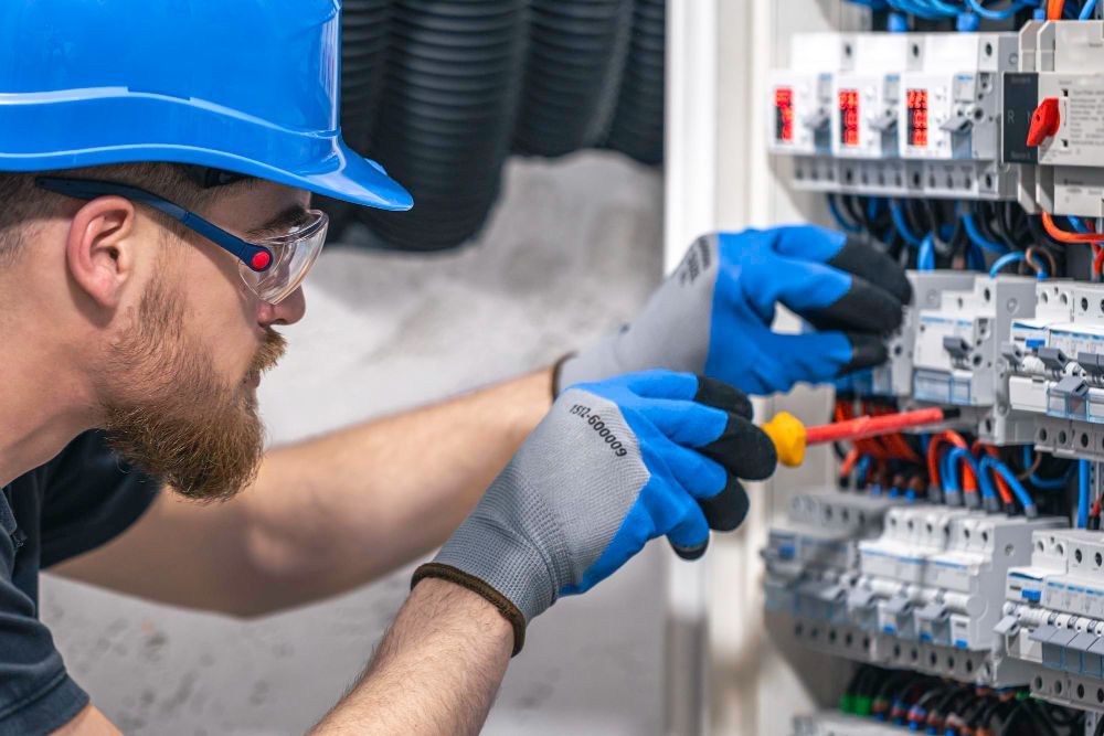A man wearing a hard hat and gloves is working on an electrical box.