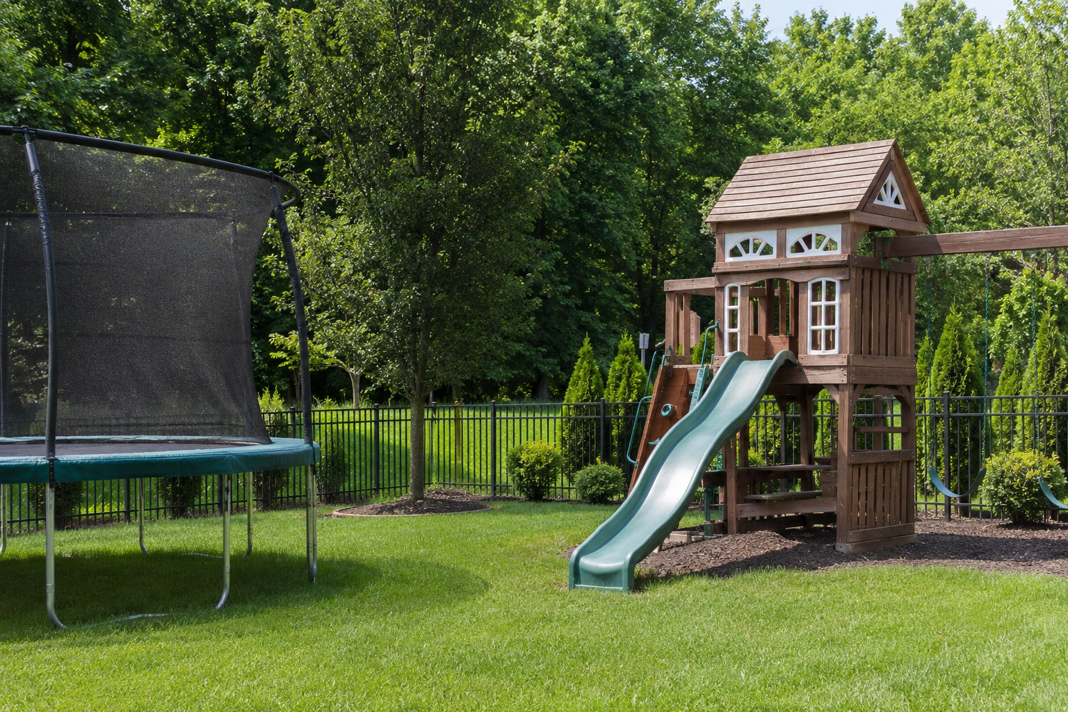 Backyard playset with a slide beside a trampoline on a green lawn, surrounded by trees.