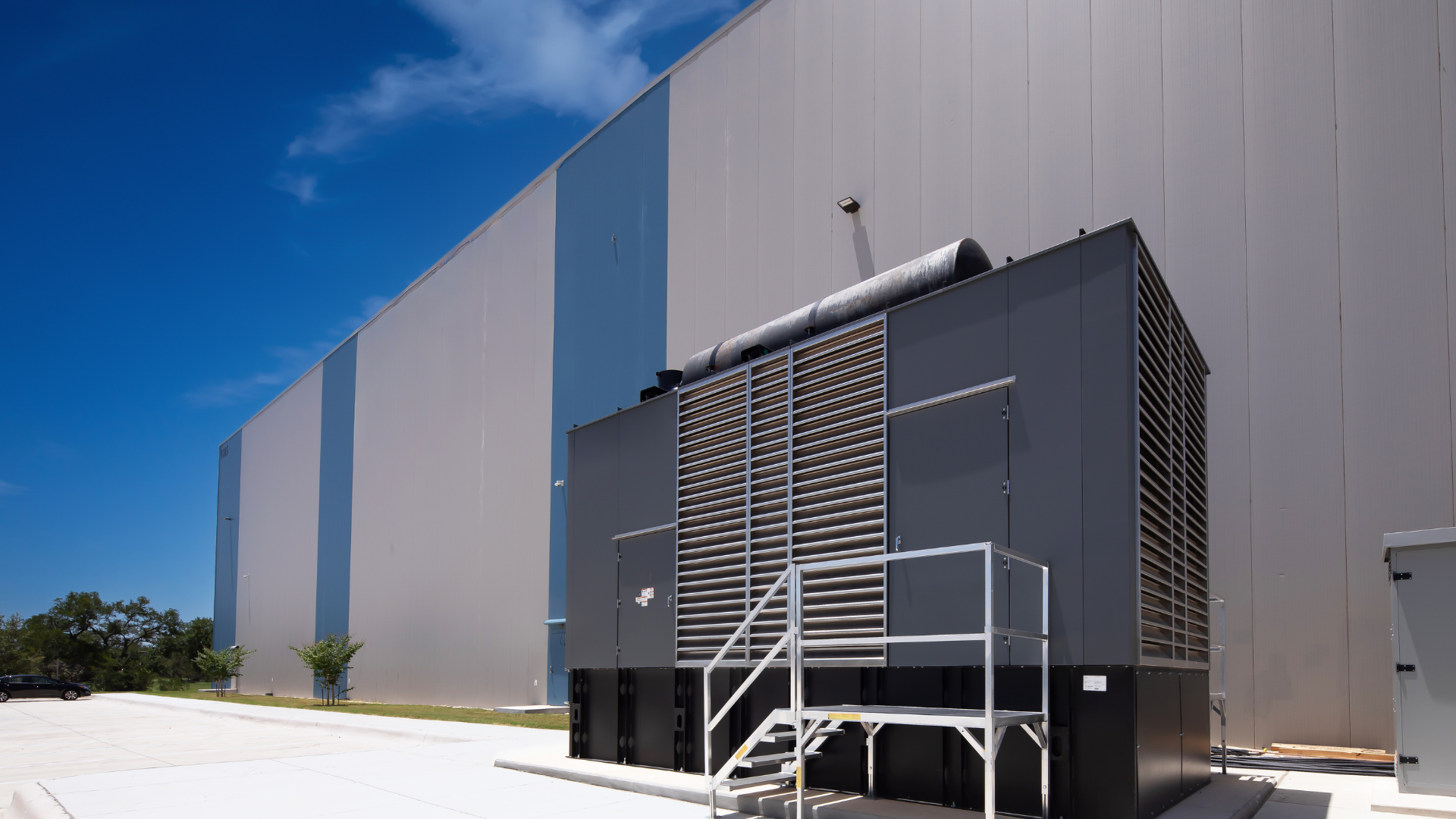 Large gray generator outside a building with light blue and gray metal siding, under a blue sky.