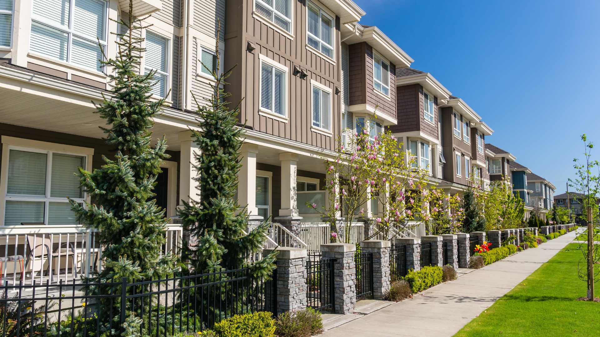 Row of modern townhouses with brown siding, a sidewalk, and green lawns on a sunny day.