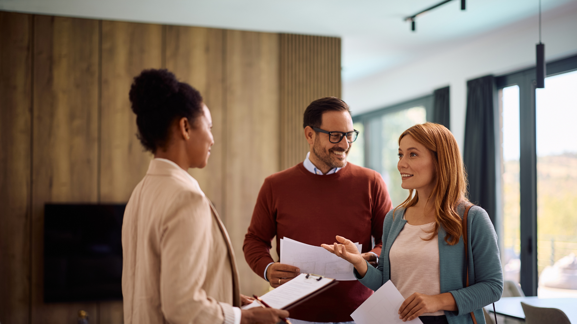 Three people stand in a modern living space, discussing papers while looking at each other.
