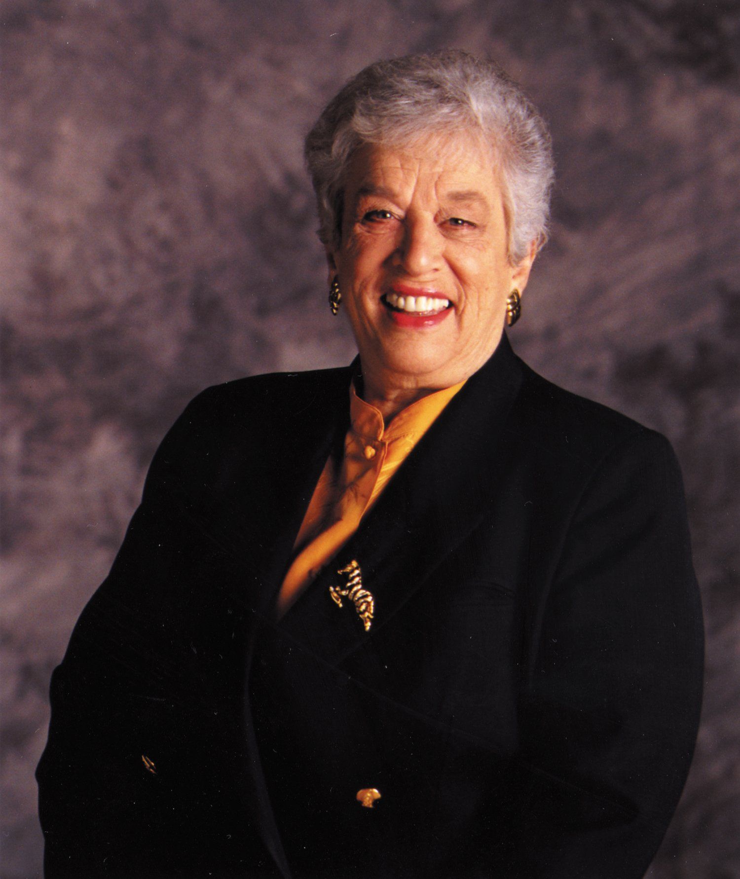 A woman wearing a black jacket and a gold brooch smiles for the camera