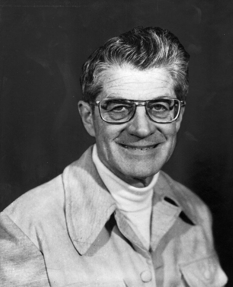 A man wearing glasses and a jacket is smiling in a black and white photo.