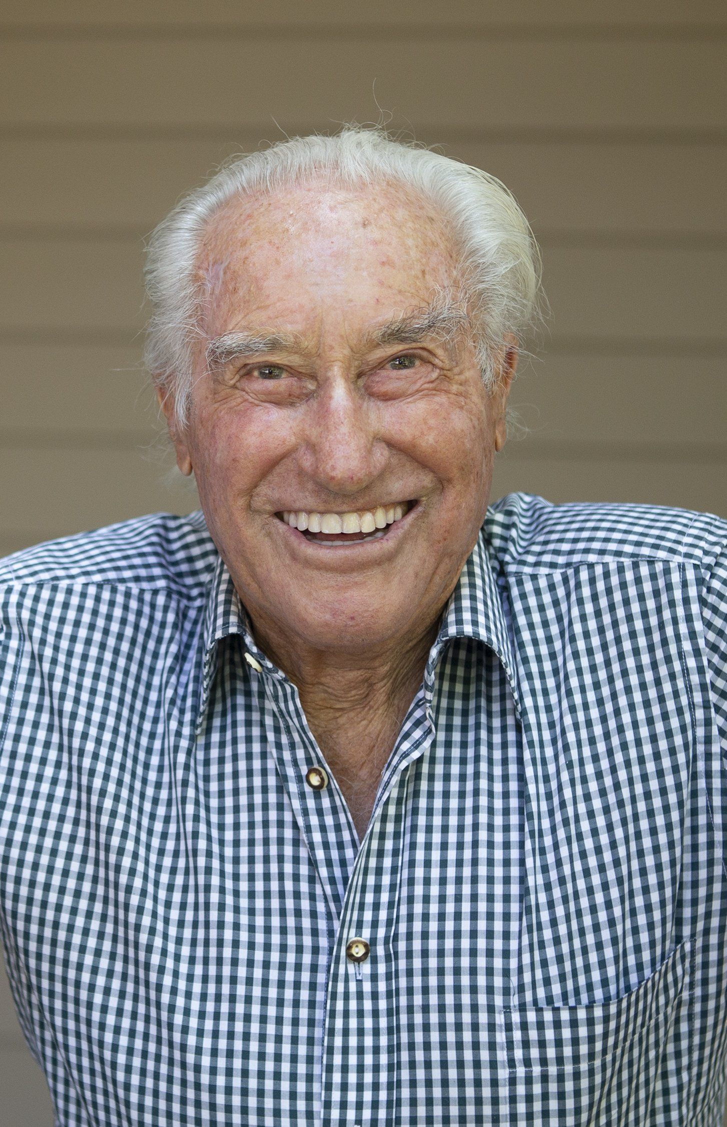 An elderly man in a plaid shirt is smiling for the camera.