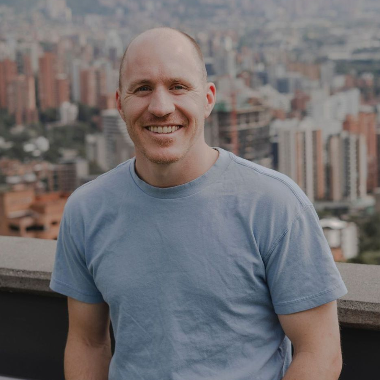 Adam Broda of Broda Coaching smiles, wearing a light blue shirt, cityscape background.