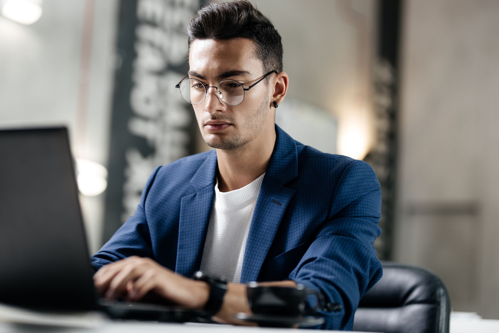 A man wearing glasses is sitting at a desk using a laptop computer, contemplating hiring a career coach.