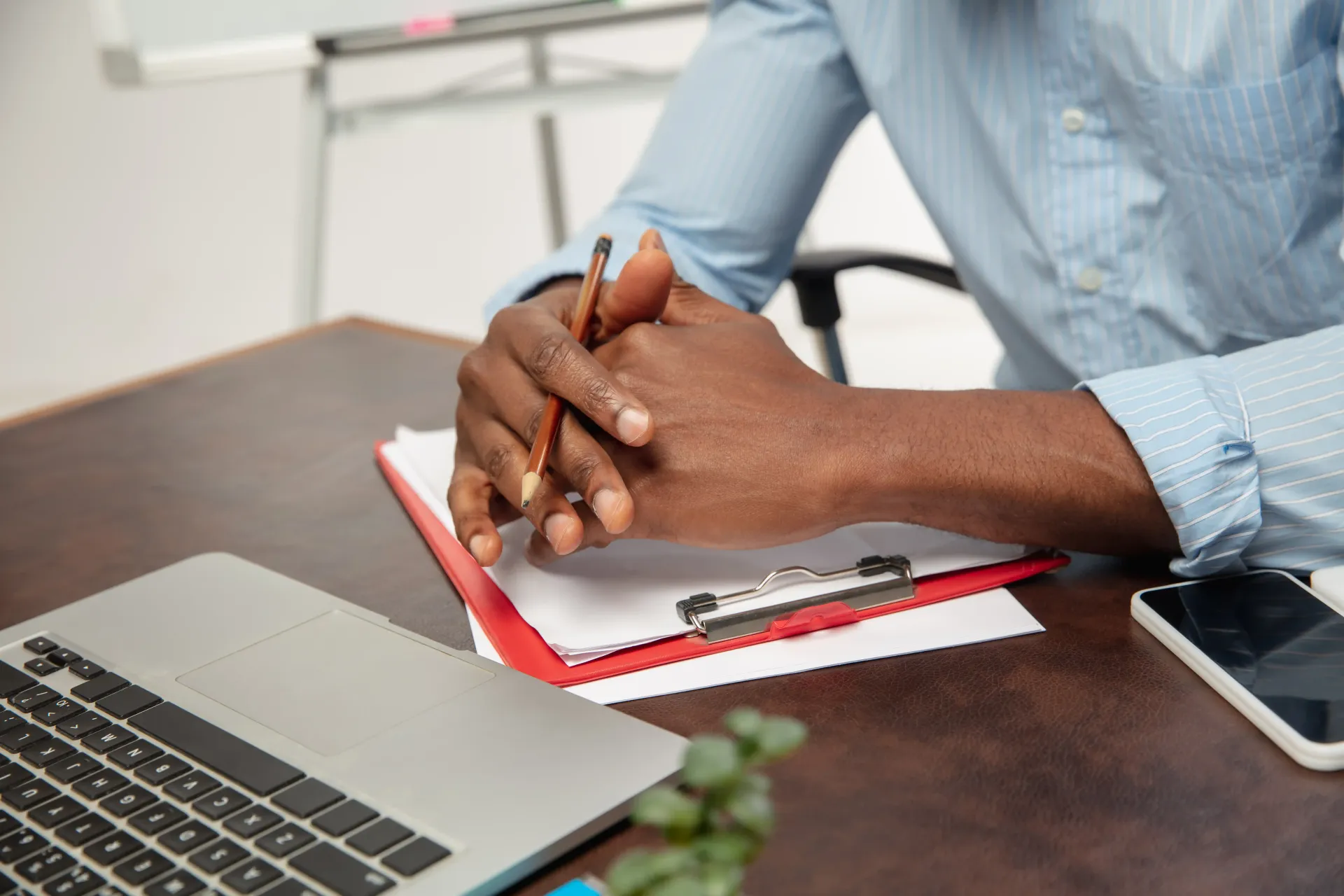Hands pencil resting on a red clipboard and desk, near a laptop and phone, applying Broda Coaching's resume writing tips.