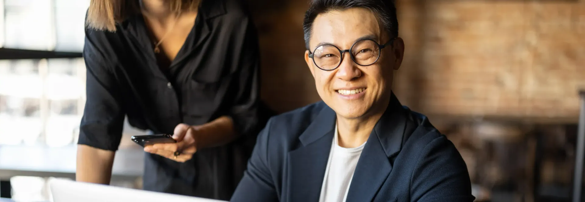 A man wearing glasses is sitting at a desk with a woman standing behind him as he works on his personal branding.