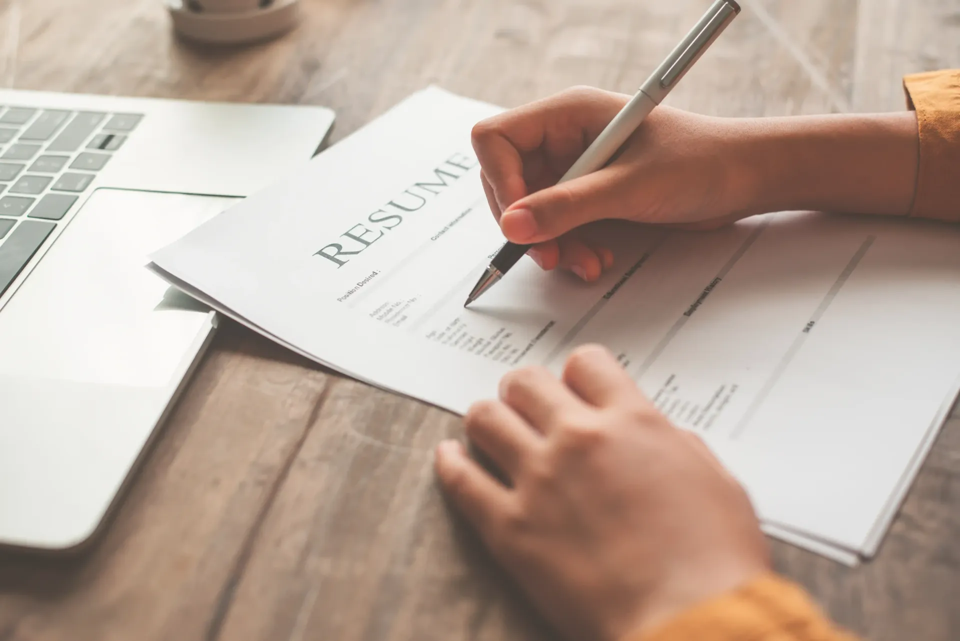 Person writing on a resume with a pen, laptop in background, following Broda Coaching's free resume template.