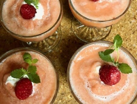 Four glasses of smoothies with raspberries and whipped cream on top.