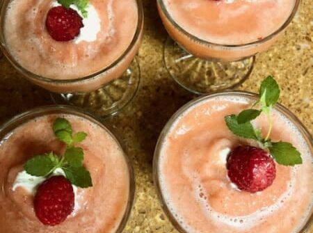 Four glasses of smoothies with raspberries and whipped cream on top.