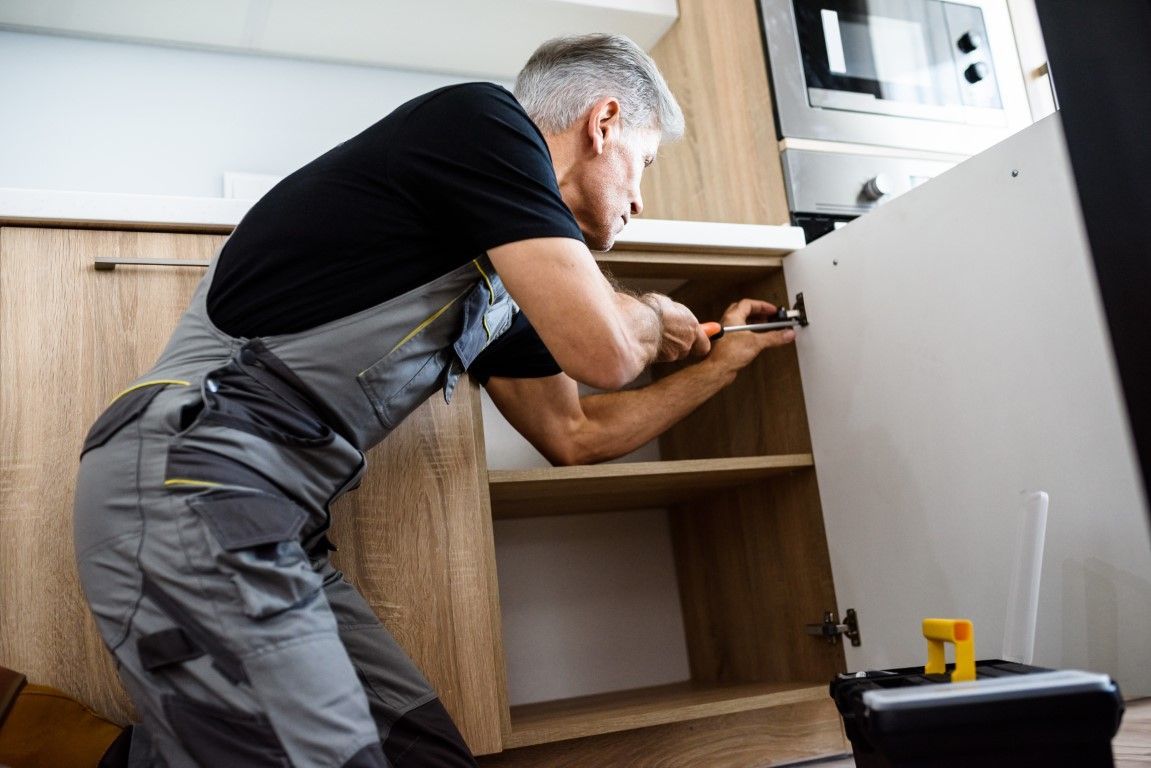 Handyman in gray jumpsuit installing a cabinet door in a kitchen.