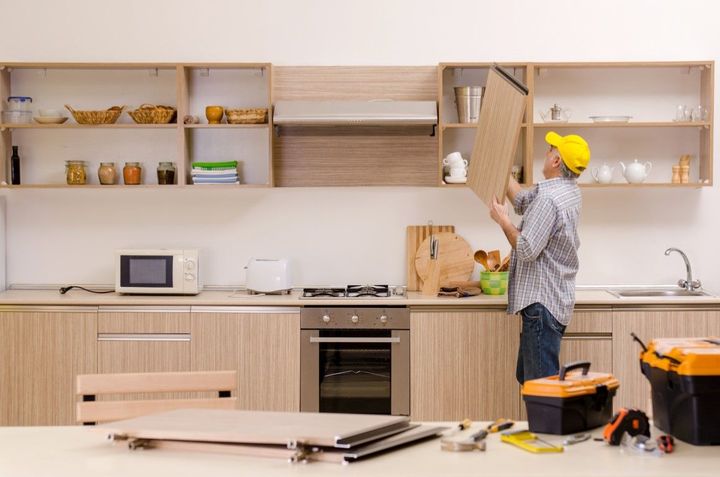 Man in yellow hard hat installing a cabinet in a kitchen with light wood cabinetry, tools, and appliances.