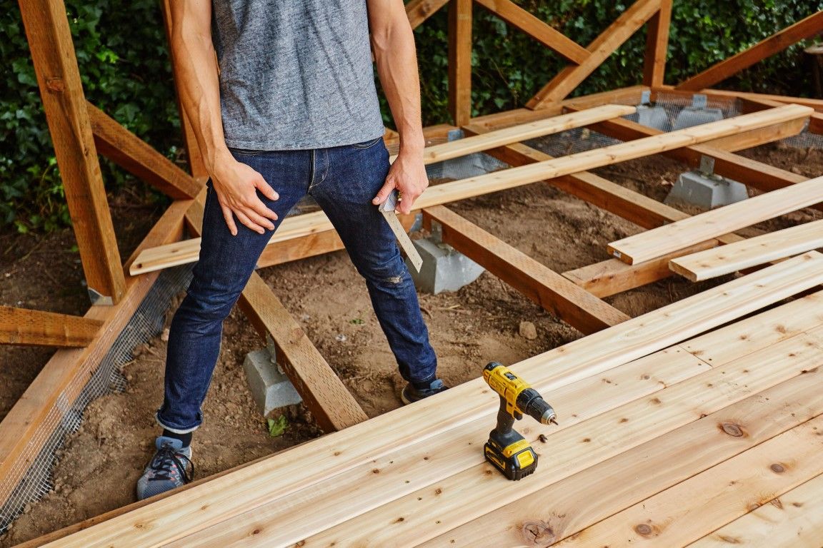 Person building wooden deck with drill, dirt ground, and wooden frame in a yard.