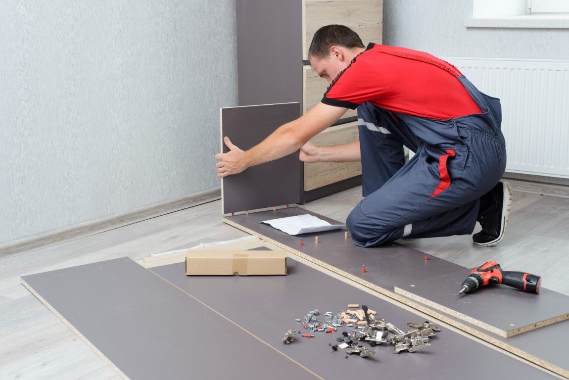 Man in red shirt and blue overalls assembling furniture in a room, using a drill.