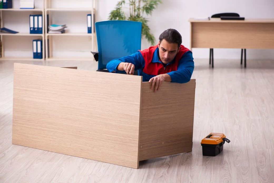 Man assembling a cardboard box in an office, with a toolbox nearby and a blue chair in the background.