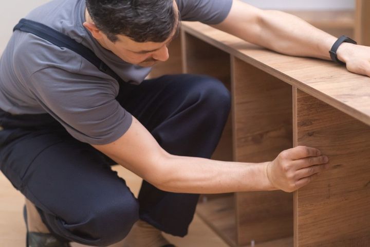 Man in overalls assembles wooden shelves, kneeling indoors.