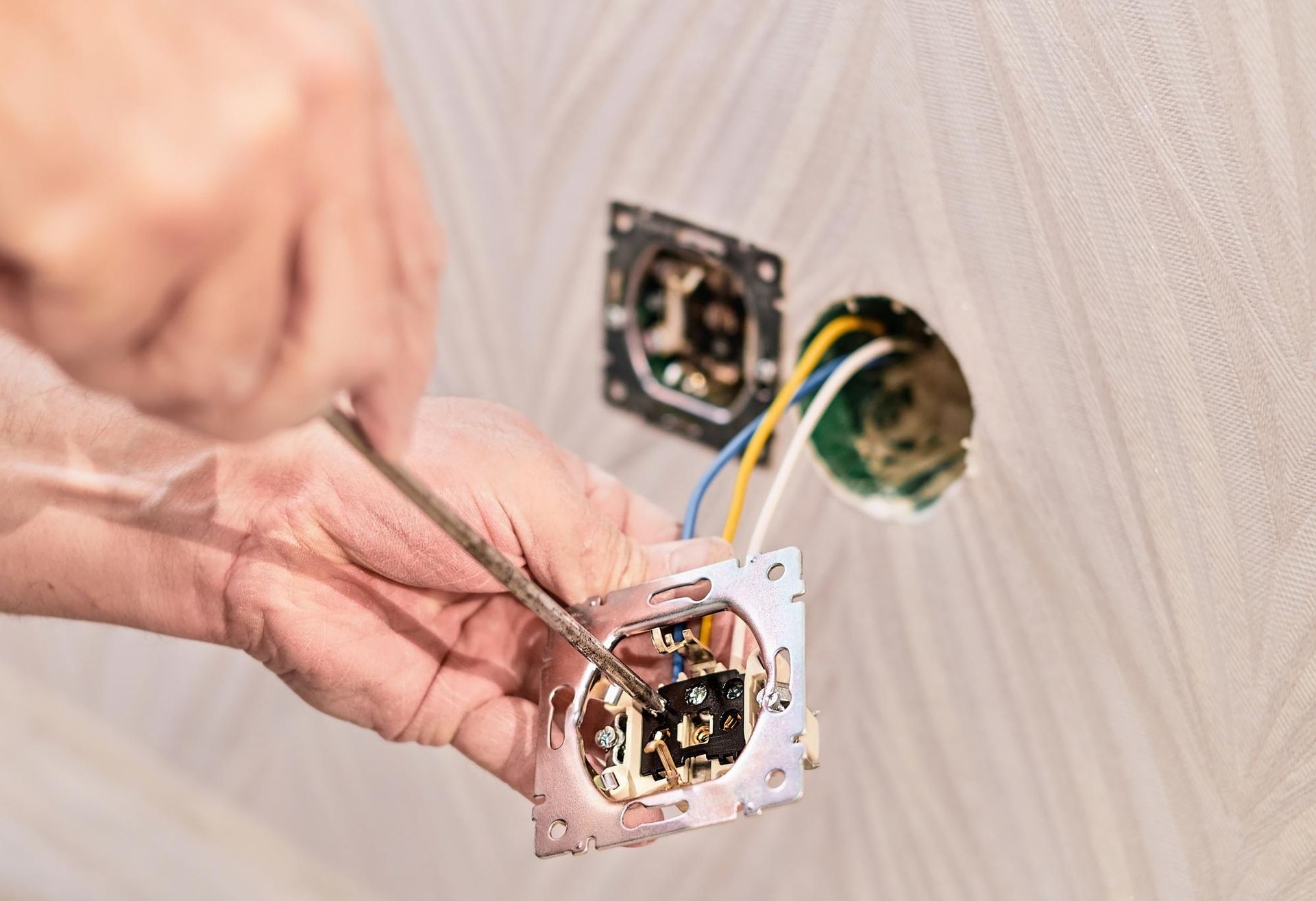 Hands wiring an electrical wall outlet box with exposed cables and tools