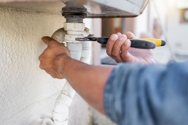 Plumber tightening a white PVC pipe under a sink with a wrench