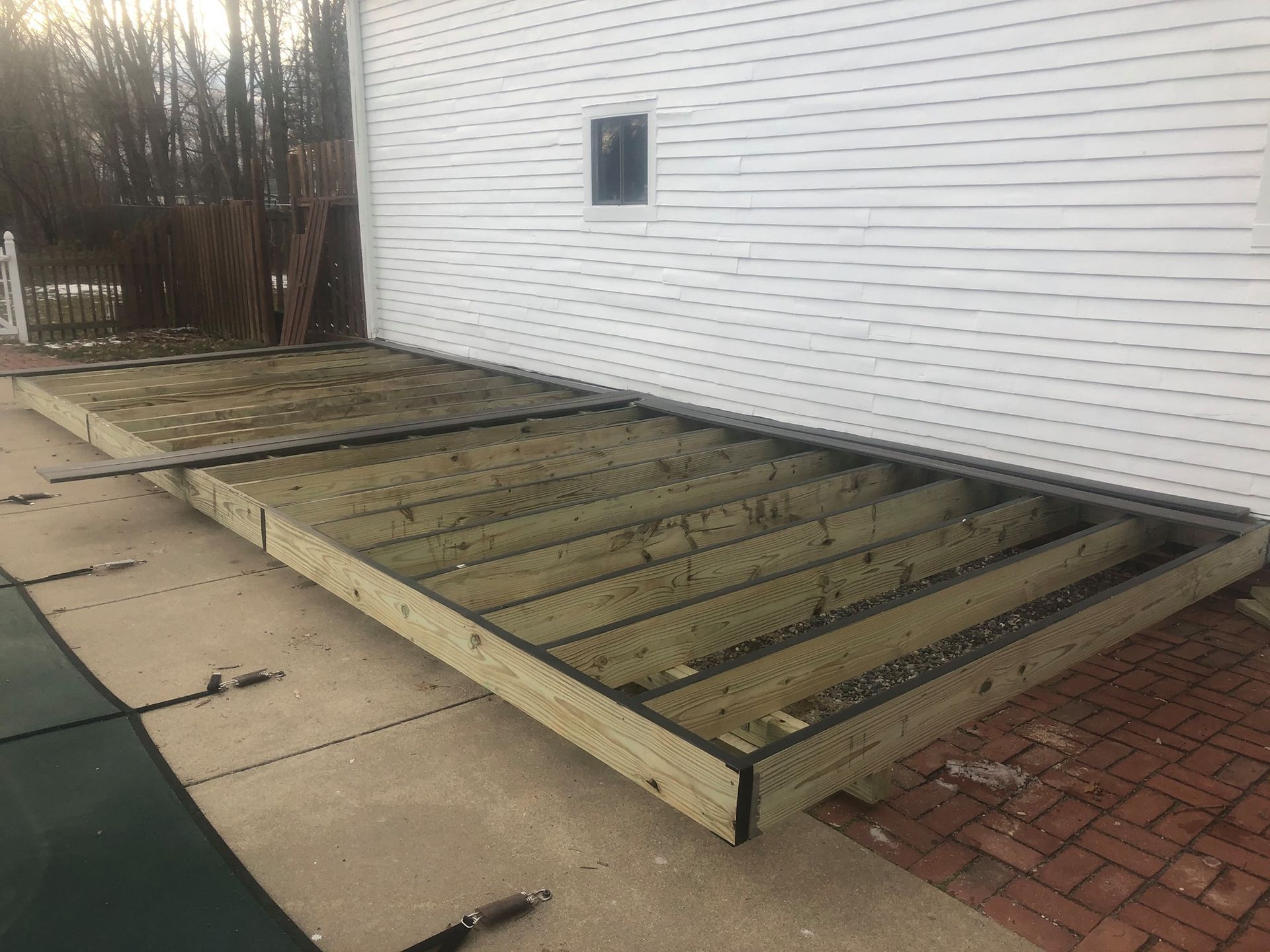 Wooden deck in a backyard with a red fence and houses in the background. Wooden deck frame next to a white house, built on a brick patio.