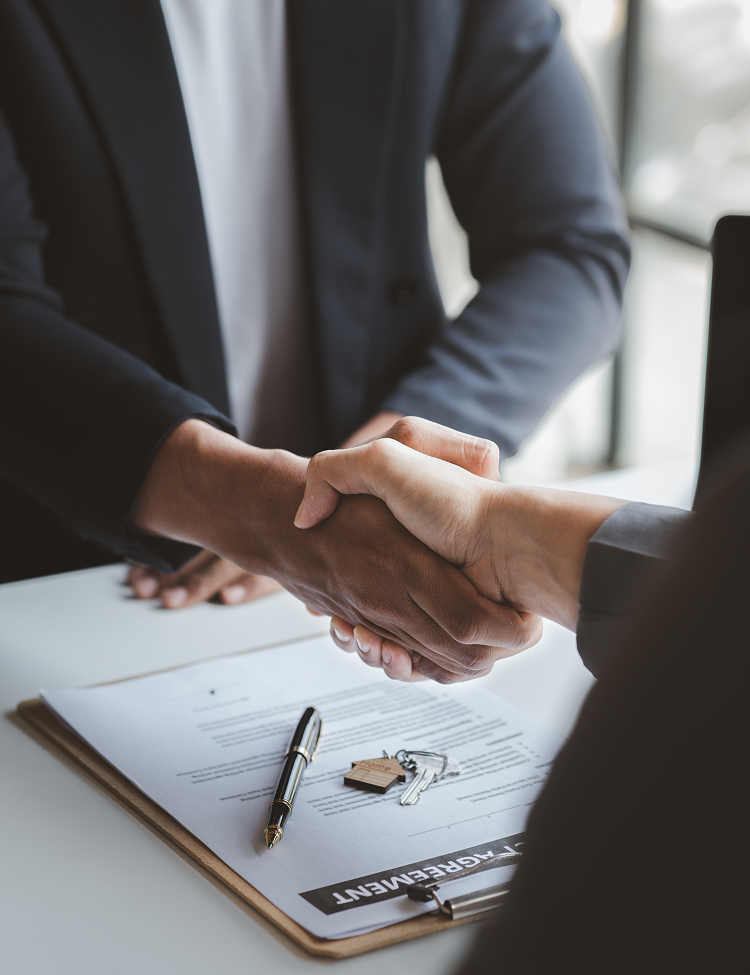 Two people in suits shaking hands over a document with keys and a pen on a table.