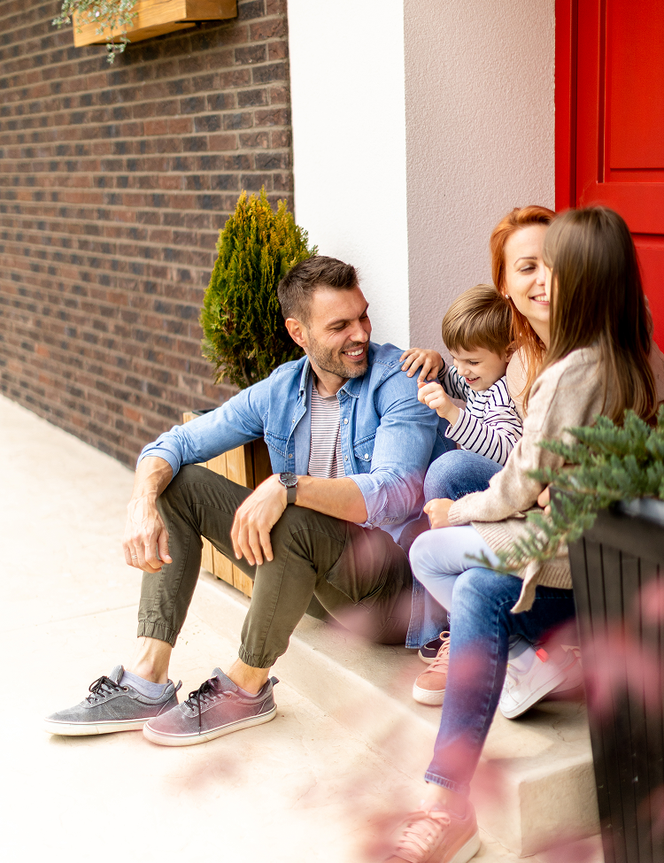 A smiling family of four sits on the porch steps of their home. The father looks toward his wife and son.
