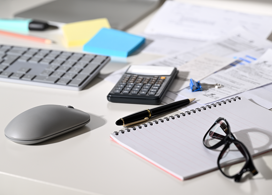Cluttered desk with a calculator, notepad, pen, keyboard, mouse, papers, sticky notes, and glasses.