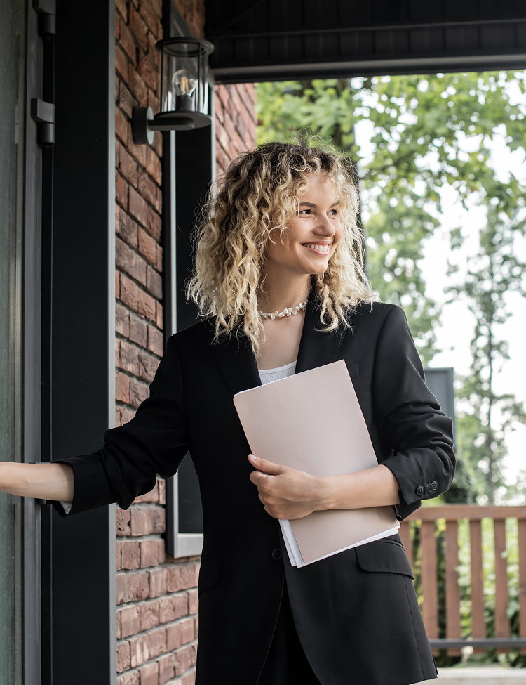 Woman with curly blonde hair, in a black blazer, smiles while holding files at a doorway. The setting is a brick building.