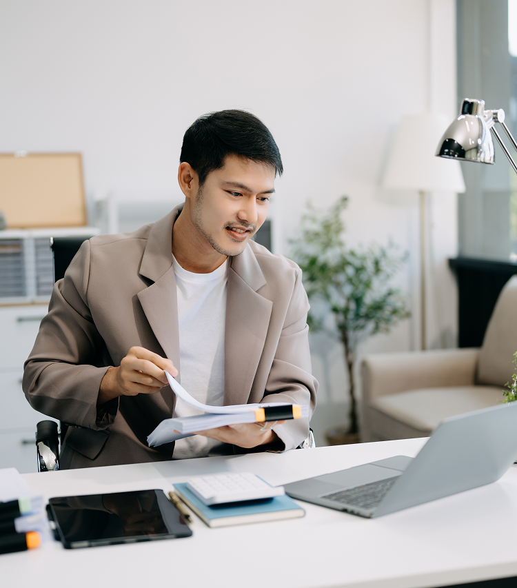 Man in blazer smiles, reviewing papers at a desk with a laptop. Bright, modern home office setting.