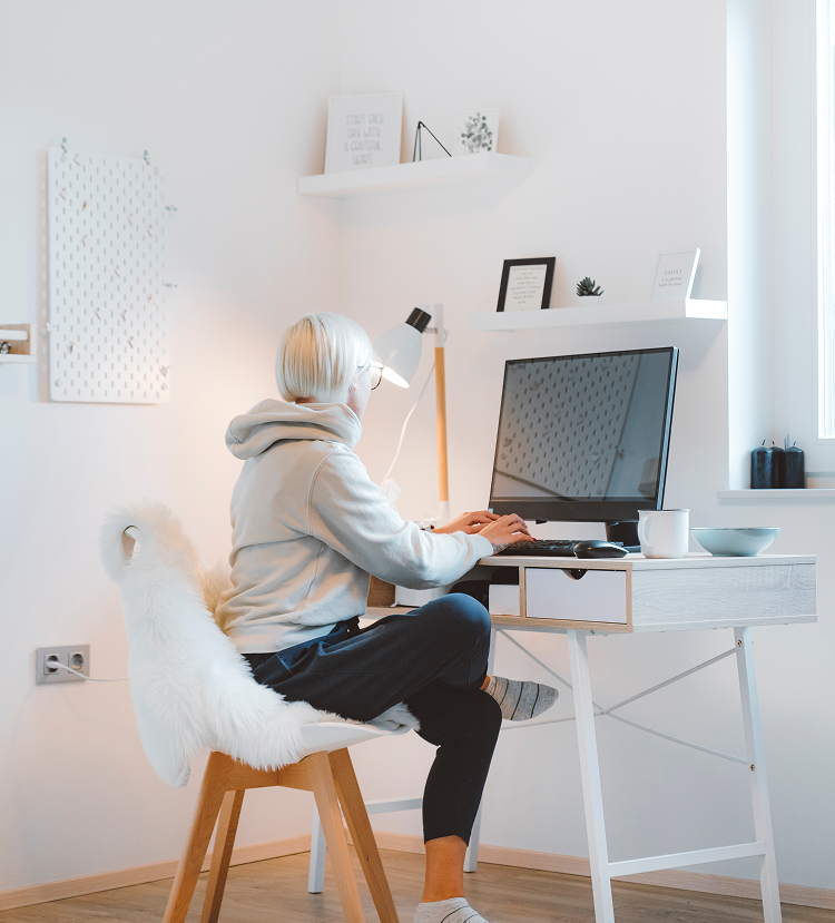 Woman in a grey hoodie working at a white desk with a computer in a bright room. She is sitting in a fluffy chair.