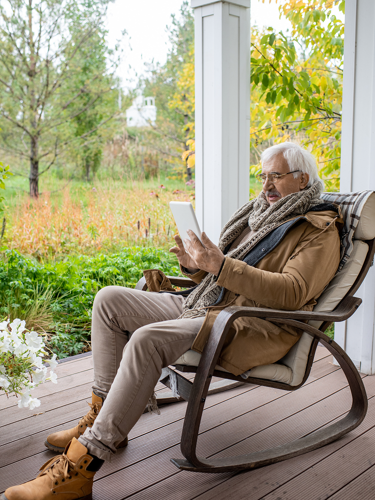 An older man with glasses reads a tablet while sitting in a rocking chair on a porch