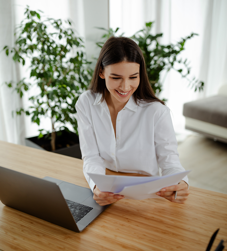 Woman smiles, reviewing documents at a wooden desk with a laptop, near a potted plant in a bright room.