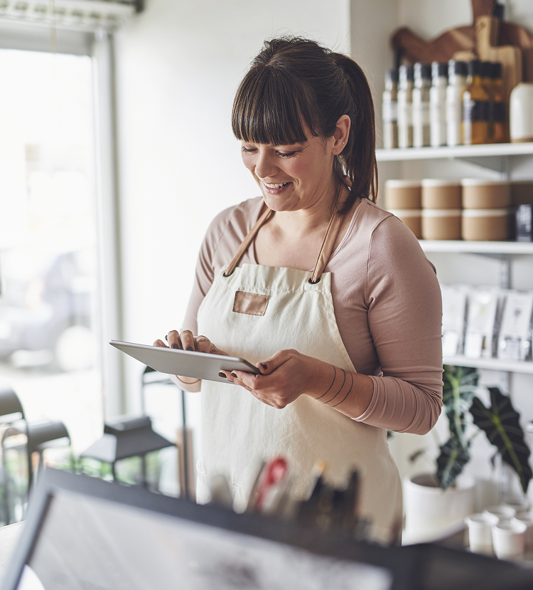 Woman in an apron smiling while using a tablet at a store counter. Bright interior with shelves of products.