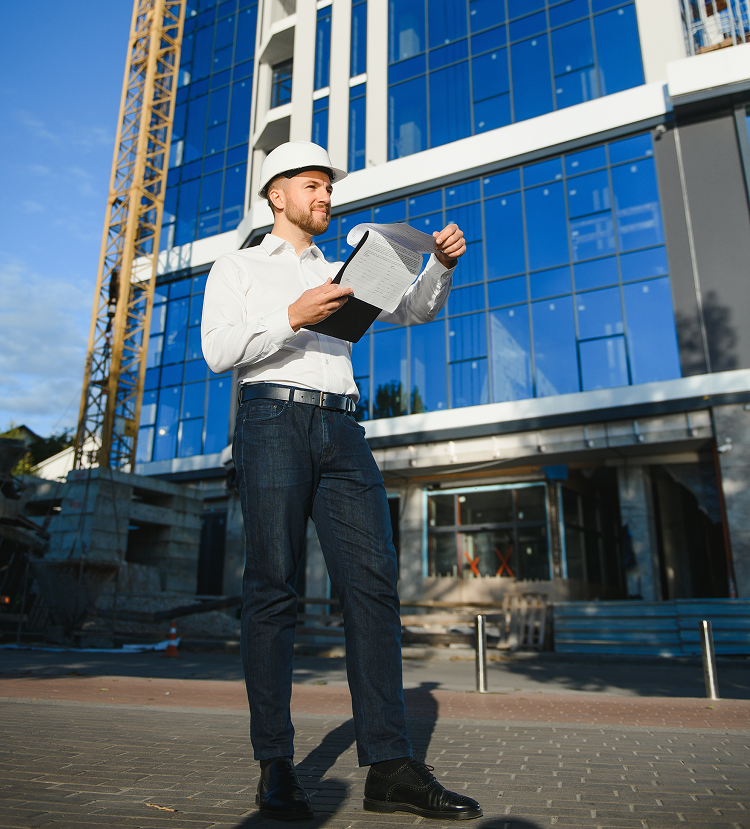 Construction worker in hard hat examines blueprints in front of a modern building under construction on a sunny day.