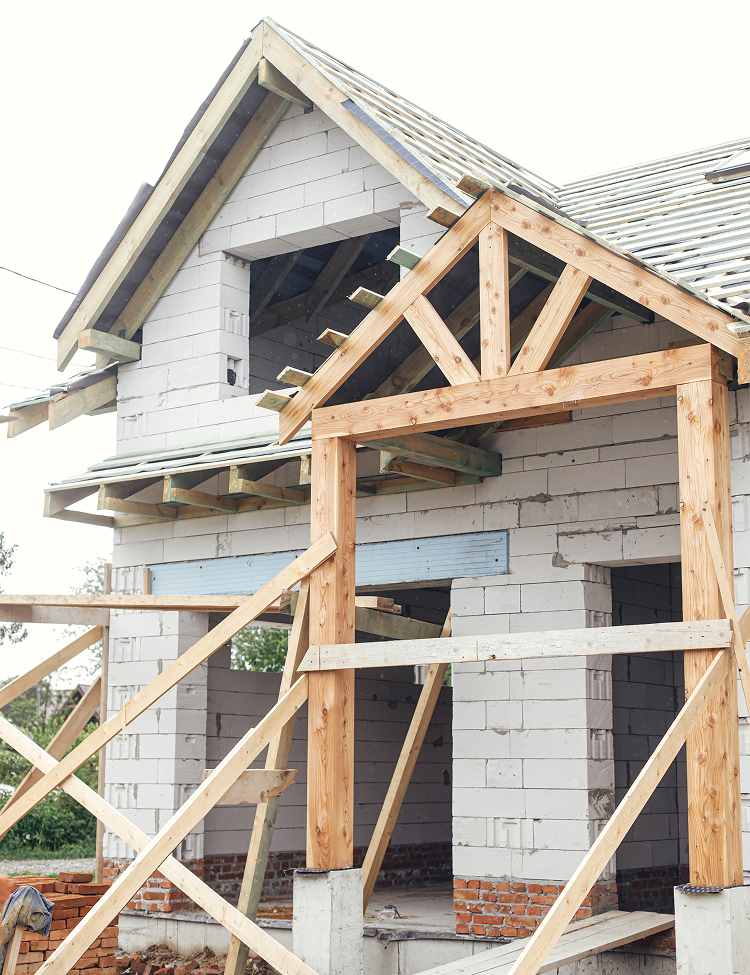 A two-story house under construction with white block walls, exposed wooden framing, and a partially built front porch.