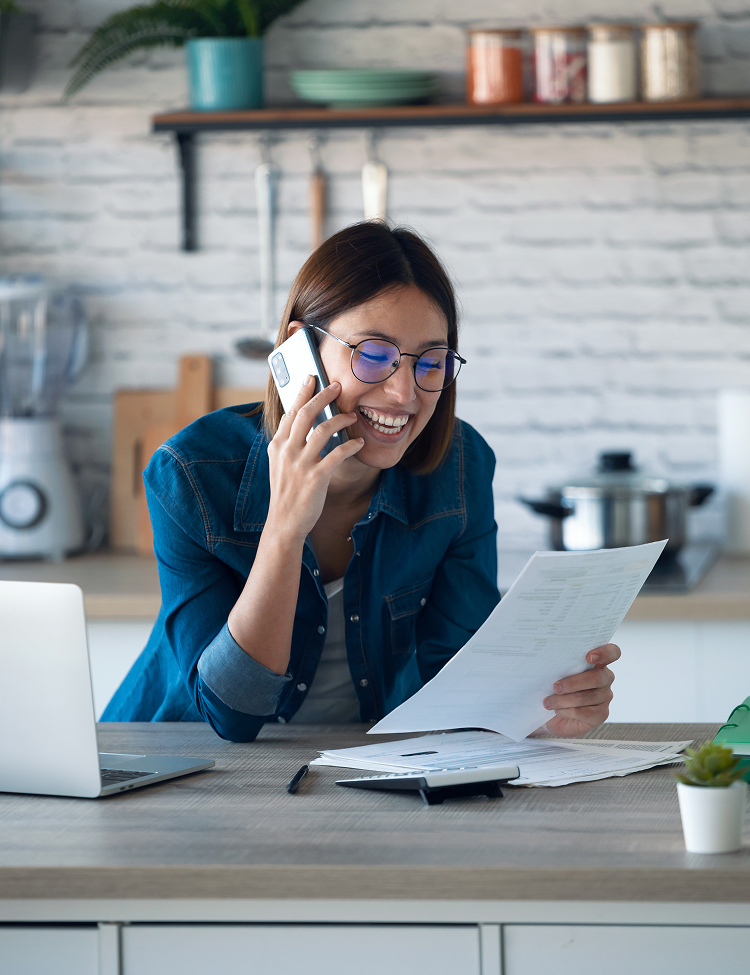 Woman in kitchen, smiling while on phone, looking at papers and laptop.
