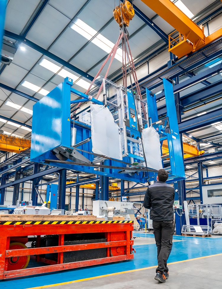 A worker watches as an overhead crane lifts a large blue and white industrial machine in a factory setting.