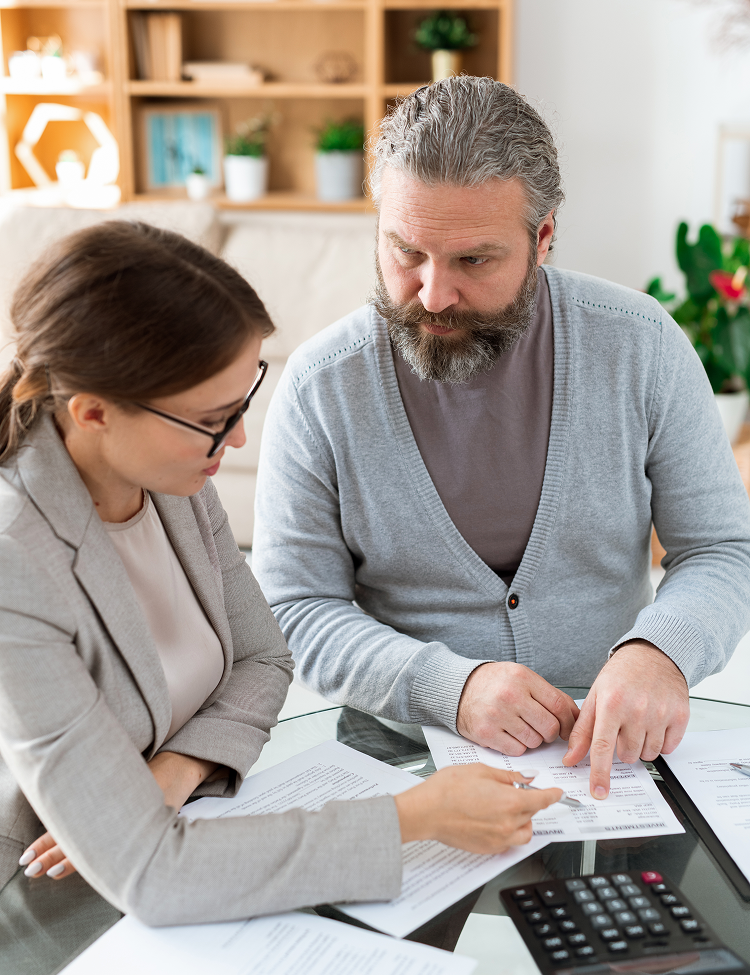 A woman in glasses points at paperwork while discussing finances with a bearded man at a table with a calculator.