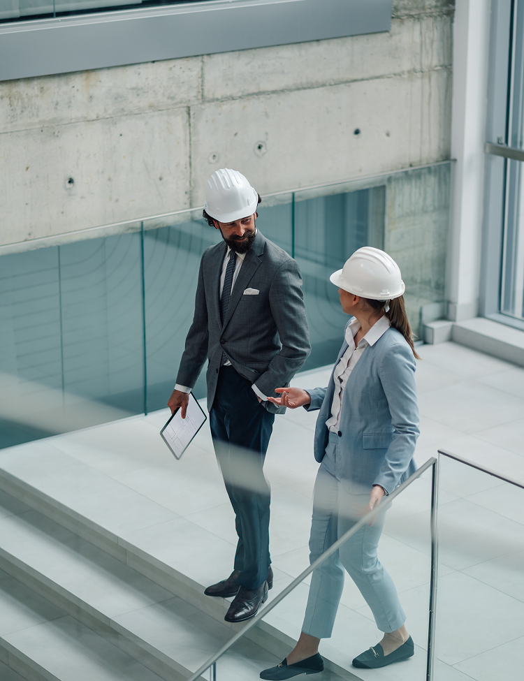 Two people in hard hats and suits walking up stairs in a modern building, discussing something.