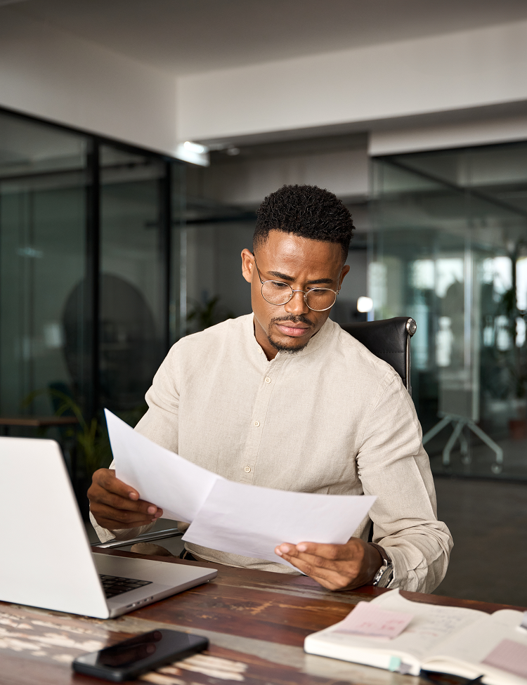 Man in glasses examines paperwork at his desk, working in a modern office setting. He has a serious expression.