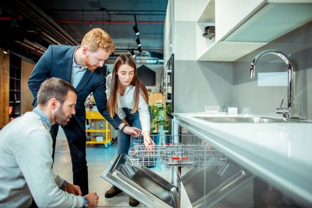 Three people examining a dishwasher in a showroom; a salesperson shows a couple the interior.