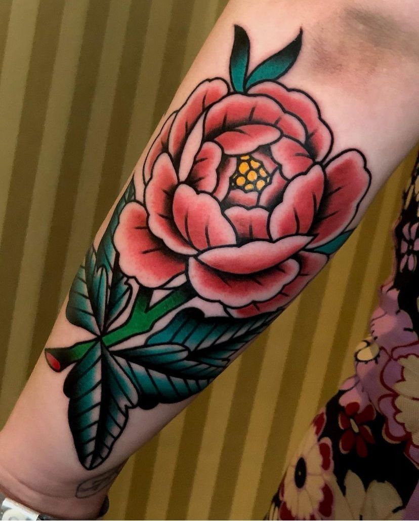 Close-up of a forearm tattoo showing a pink peony with green leaves