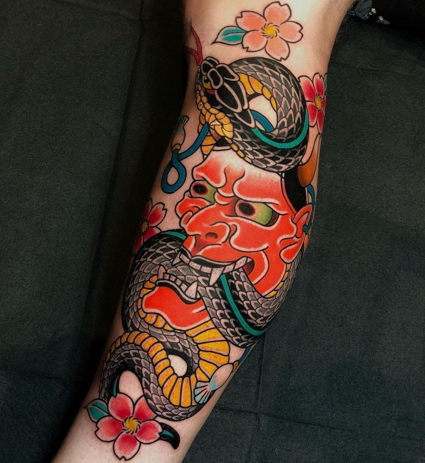 Colorful Japanese-style tattoo on a forearm with a red face, koi fish, snakes, and pink flowers