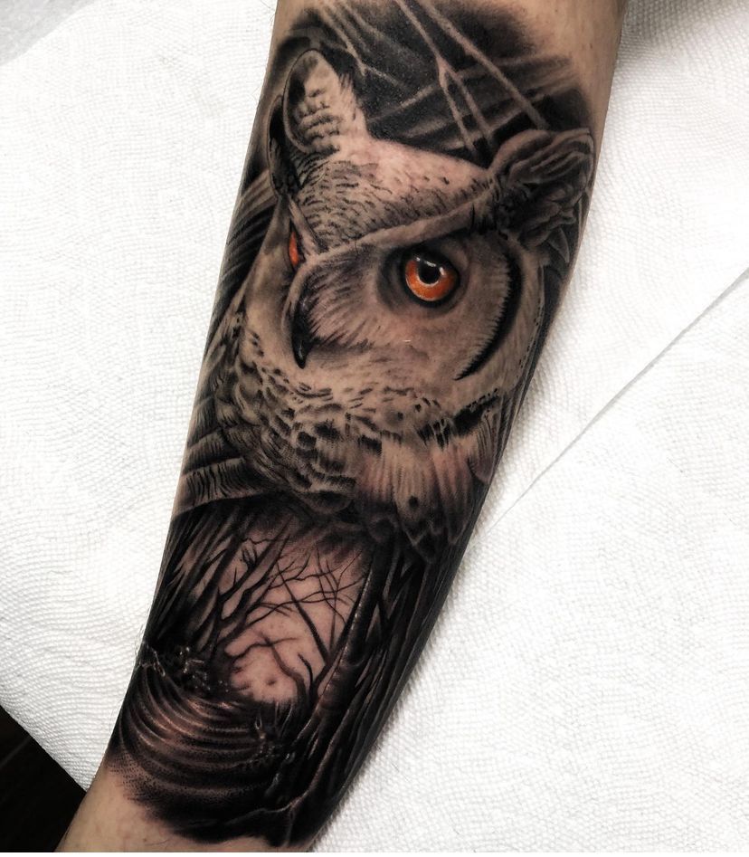 Detailed owl tattoo on a forearm, with a striking orange eye and dark feathered shading