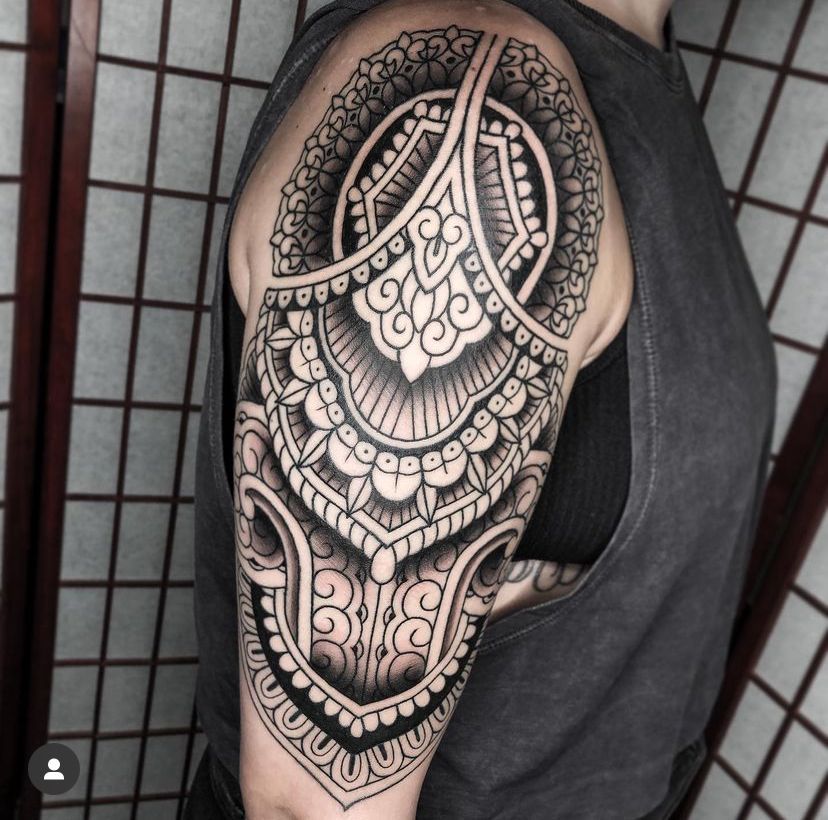 Black-and-gray ornate tattoo covering upper arm and shoulder, shown in profile on a person wearing a dark sleeveless top