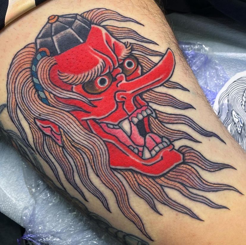 Red demon mask tattoo with black hair and flames on a person’s arm