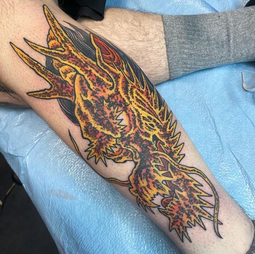 Colorful dragon tattoo on a leg, with orange, yellow, and black flames on a shaded blue background