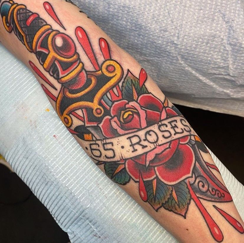 Colorful tattoo of a red rose and banner reading “55: ROSE” on a forearm